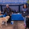 Two volunteers from the Exeter group stand by a stall with Guide Dogs merchandise and two dogs.