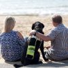 A guide dog sits on the beach in between two people. He has a yellow and white harness on.