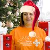 woman wearing Santa hat and orange T-shirt with gifts standing in front of a Christmas tree
