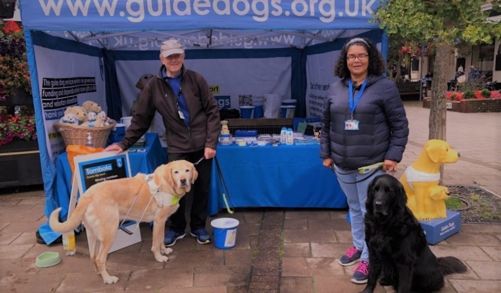 Two volunteers from the Exeter group stand by a stall with Guide Dogs merchandise and two dogs.