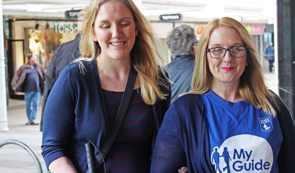 Two women walking along smiling, one provides on arm guiding to the other who is holding a long cane