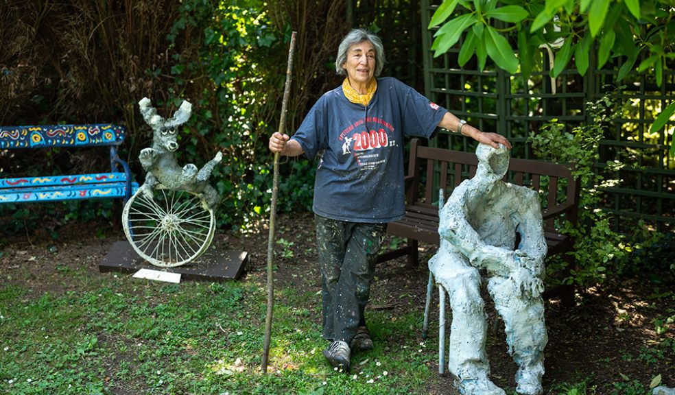 woman with grey hair holding a stick with other hand resting on the head of a sculpture