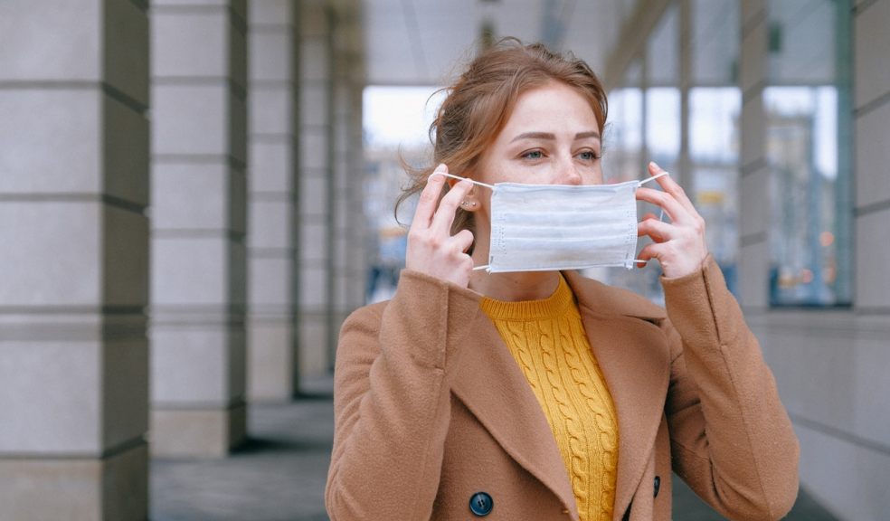 Woman in a brown jacket and yellow jumper placing a disposable face mask on her face