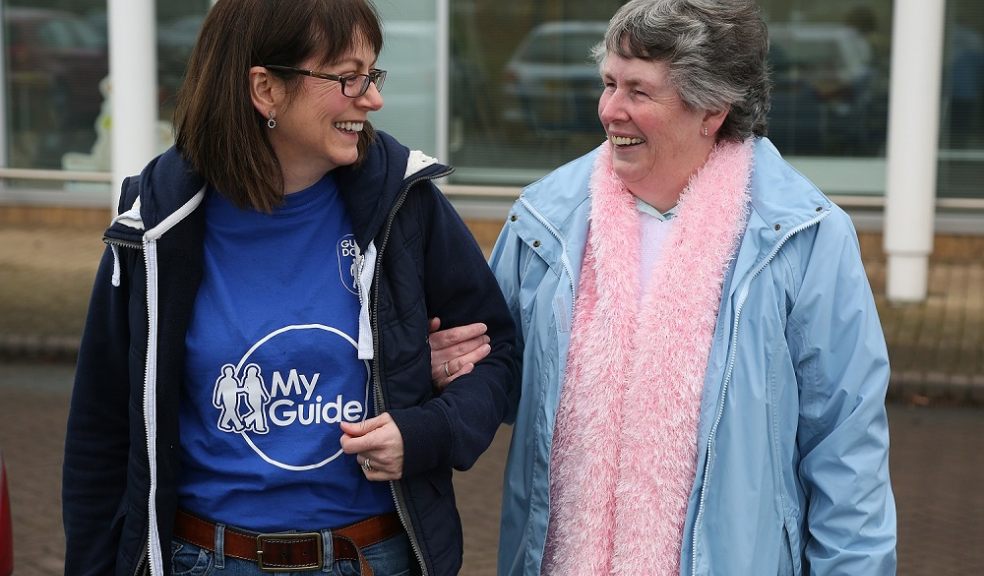 two women walking together chatting and laughing, one provides on arm guiding, the other is visually impaired