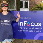 Photo is of an 11 year old girl, wearing a blind fold, standing in front of InFocus Charity Logo