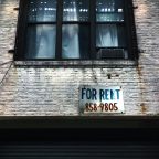A weathered for rent sign fixed to the wall below a window on a city building