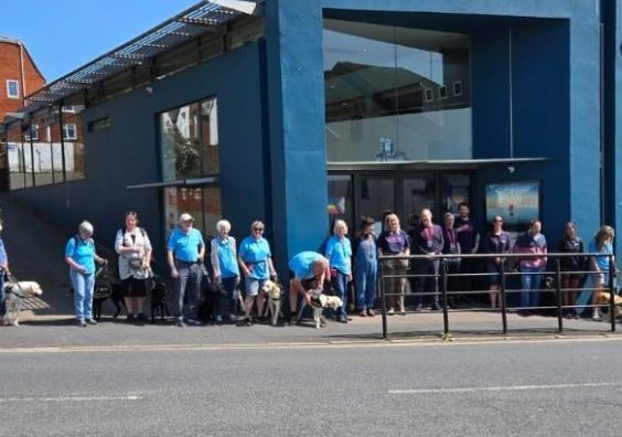 A group of volunteers with their guide dog pups in training stand in front of the Exeter Picturehous