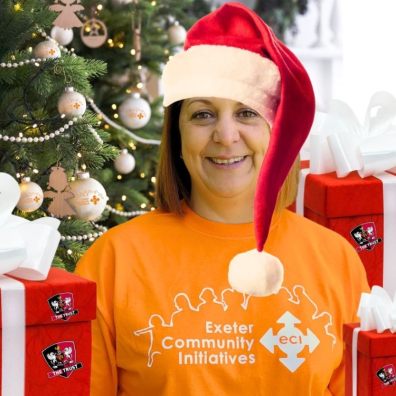 woman wearing Santa hat and orange T-shirt with gifts standing in front of a Christmas tree
