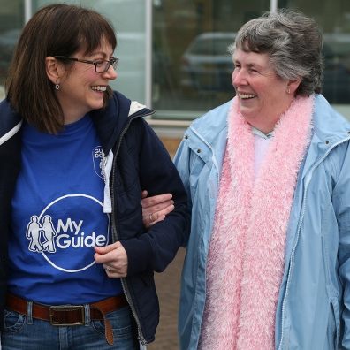two women walking together chatting and laughing, one provides on arm guiding, the other is visually impaired